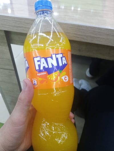 A clear plastic bottle of Fanta Apelsin (orange flavor) is held in a human right hand. The bottle contains a vibrant orange-yellow carbonated beverage and is sealed with a blue screw cap. An orange label encircles the bottle, displaying the white "FANTA" logo, the word "Apelsin," an illustration of an orange slice with liquid splashes, and text indicating "80 kcal" per "250 ml" and a "Vitamin C" symbol. The background consists of a light-colored, reflective surface, consistent with a wooden or laminate countertop, positioned in the upper right. Below this surface, a dark, blurred area reveals indistinct shapes suggestive of white footwear and dark clothing. The scene is set indoors, in Gijduvan, Uzbekistan.