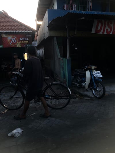 The image depicts an outdoor scene, likely in Ngaban, East Java, with a sense of casual activity. Two individuals are present. One person, with their back to the camera, is wearing a dark t-shirt and shorts, with dark sandals. Their legs and feet are visible as they stand near a bicycle, with one foot resting on a pedal. The other person is partially visible in the background, shirtless and wearing a cap.
The scene is set on a brick-paved ground. Several objects are prominent: a bicycle with a black frame and spoked wheels is in the foreground, and a motorcycle, painted blue and white, is parked behind it. The motorcycle has a license plate and a sign that reads "DIJUAL" (for sale) and a phone number.
In the background, there are structures that appear to be part of a building or shop. A large, red and white sign with the text "DIS" is visible on one of the buildings. Further to the left, another sign with red text and the number "76" is partially visible, along with the words "DIJARUM" and "APEL ROYAL BARU".
The lighting suggests it is either early morning or late afternoon, with soft, directional light casting shadows. The overall mood seems relaxed and everyday, with no indication of a specific event or strong emotion. The location cues, such as the signs in Indonesian and the architectural style, strongly suggest an Indonesian setting.