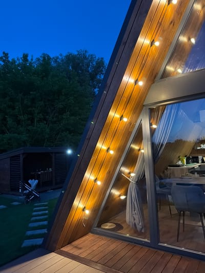 This is a nighttime exterior shot of a modern A-frame house in Sharovechka, Ukraine. The house has a wooden exterior and a large glass window. String lights are hung along the wooden exterior. Inside the house, a dining table with chairs is visible through the window. In the yard, there is a gazebo with a table, a stroller, and a pathway made of flat stones. The sky is dark blue, indicating it is dusk or night.
