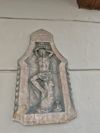 This is a close-up, eye-level, outdoor shot of a weathered stone relief sculpture mounted on a light-colored, textured wall. The sculpture is set within an arched, niche-like frame with a twisted rope motif along the sides. The main subject of the relief is a figure of Saint Sebastian, depicted bound and pierced by arrows. His body is contorted, and his hands are raised behind his head. He is wearing a loincloth. At the bottom of the relief, to the right of the figure's feet, is a small relief of a bell.
Above the figure of Saint Sebastian, carved into the stone, is an inscription in German. The visible text reads: "ST. SEBASTIAN / von krankheit breil / unaung erbolent / und schmerzel / im letzlen End." This translates to "ST. SEBASTIAN / from illness free / healing entreats / and suffering / in the end."
The sculpture appears to be quite old, with signs of wear and erosion, particularly on the figure and the inscription. The stone itself is a pale pinkish-grey, with some darker grey and reddish-brown discolorations. The wall behind the sculpture is painted a light, creamy white and has a slightly bumpy, uneven texture. A thin, dark horizontal line is visible across the wall above the sculpture, possibly indicating a seam or a change in building material. In the upper right corner of the image, a sliver of wooden structure, possibly eaves or a roof beam, is visible. The lighting suggests it is daytime, with diffused light. The setting appears to be an exterior wall of a building, likely in a historical European town, given the context of Kitzbühel, Austria.
