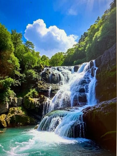 The image depicts a majestic waterfall cascading down a rocky incline, surrounded by lush green trees and a vibrant blue sky with fluffy white clouds. The water is a striking turquoise color as it collects in a pool at the base of the falls, creating a serene and natural landscape. The rocks are mossy and weathered, suggesting a humid and ancient environment. The scene is devoid of any human presence, focusing entirely on the raw beauty of nature. The location specified, Konstantinovka, Kyrgyzstan, is not discernible from the image itself, as it is a natural landscape and not within an urban setting.