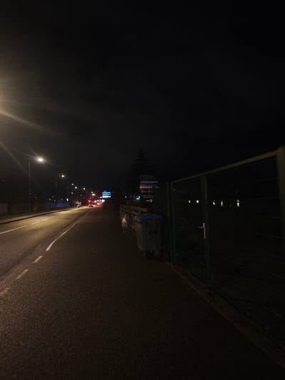 A nighttime street scene shows a dark road illuminated by streetlights leading towards distant car lights. A sidewalk runs along the right, bordered by a green chain-link fence. Adjacent to the fence are overflowing trash bins and a signpost with business information. The sky is dark and overcast, adding to the somber, urban atmosphere.
