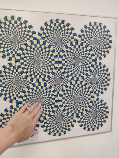 The image shows a framed optical illusion artwork featuring a repeating pattern of concentric circles with alternating blue, yellow, and black shapes. A person's left hand, with polished nails and rings, is reaching out to touch the artwork, with the fingertips making contact with one of the circular designs. The artwork is displayed on a white wall. The context provided indicates this scene is located in Thessaloniki, Greece.