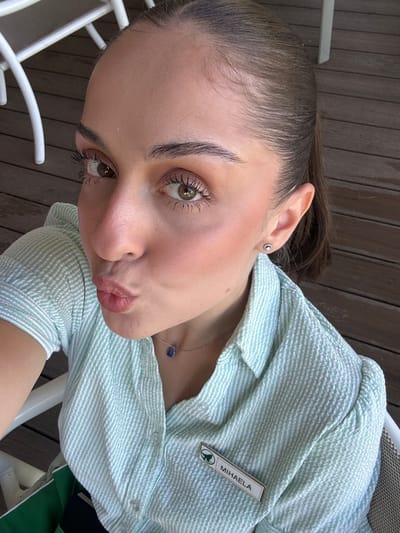 A young woman with her hair pulled back in a ponytail, wearing a mint green and white striped collared shirt and a name tag that reads "MIHAELA," leans in towards the camera, puckering her lips. She has on subtle makeup, including mascara, eyeshadow, and blush, and is wearing a delicate necklace with a blue pendant and small stud earrings. The background suggests an outdoor setting with a wooden deck and white outdoor furniture. The overall impression is a casual, selfie-style photograph.