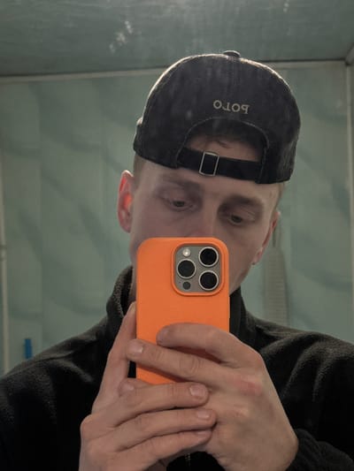 An indoor scene in Лабжанскі сельскі Савет, Belarus, depicts a light-skinned individual, likely male, taking a mirror selfie. The subject is wearing a dark, possibly corduroy, baseball cap reversed, with the mirrored word "POLO" embroidered in light thread on the back. A dark fleece jacket or top covers the torso. The subject's face is partially obscured by an orange-cased smartphone, identified as an iPhone due to its prominent triple camera array and flash on the rear. The subject's gaze is directed downwards towards the phone. The background behind the subject's reflection is a light blue-green wall, possibly tiled or painted, exhibiting minor smudges and reflections. A faint horizontal line is visible across the upper-mid section of the background, and a small, blurred vertical blue object is present in the far-left background. The individual's hands, with short fingernails, are holding the phone.