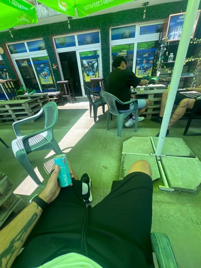 The scene depicts an outdoor patio area in Касапите, Bulgaria, under a green canopy that casts a green tint over the concrete floor. In the foreground, a person is seated, wearing black shorts, black shoes, and a smartwatch on their left arm, which bears a tattoo. This individual holds an open light blue "Red Bull THE ICED EDITION" can. To the left, a second person is seated in a green plastic chair, facing away, wearing a black t-shirt, light blue shorts, white socks, and white sneakers. They are positioned at a low table constructed from wooden pallets, on which a glass containing a yellow liquid is visible. To the right, a third person's right leg, also tattooed, is partially visible, clad in black shorts and black shoes, seated at the same pallet table. A white pole, likely supporting the canopy, stands anchored by four concrete slabs on the ground. The background features a building with windows and an entrance, adorned with artificial green foliage. Advertisements for "inbet," a betting company, are prominently displayed on posters within and around the windows, featuring sports themes such as basketball and soccer, with phrases like "BET BUILDER" and "ЕКСКЛУЗИВНО" (exclusive) in Cyrillic. A white lantern and some draped artificial vines are visible on a shelf unit behind the seated individuals. Sunlight filters through the canopy, creating distinct shadows on the ground.