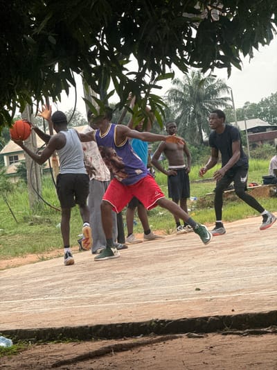 In Amansee, Nigeria, several young men are engaged in a basketball game on an outdoor court. The court appears to be made of concrete with painted lines. The surrounding environment is somewhat overgrown with grass and vegetation, and palm trees are visible in the background, suggesting a tropical or subtropical climate.
In the foreground, a young man in a purple and yellow basketball jersey with an image of Kobe Bryant is actively playing. He is wearing red shorts and green and white basketball shoes. He is in motion, possibly dribbling or passing the ball. To his left, another young man in a grey tank top and black shorts, holding an orange basketball, is also in a playing stance. He is wearing black and white athletic shoes.
Behind them, other individuals are observing or participating in the game. One young man, shirtless and wearing black shorts, stands with his hands on his hips, looking towards the players. Another young man in a black t-shirt and black leggings, with black and grey basketball shoes, is also actively involved in the game, facing the players. Further in the background, buildings and more foliage can be seen. The lighting suggests it is daytime, possibly overcast. The overall scene depicts a casual basketball game taking place in a community setting.