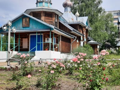 In the city of Zelenogorsk, Russia, this image captures a wooden Orthodox church with a blue-painted facade and a distinctive scalloped trim under its roof eaves. The church features multiple domes, one copper-colored and two smaller silver ones topped with crosses. A traditional icon is displayed on the front gable. In front of the church, a white railing encloses a set of stairs leading to a blue double door entrance. A modern streetlamp with three white globes stands to the left. The foreground is filled with a lush garden of blooming pink and red roses, with green foliage and a patchy grassy ground. Behind the church, a dense canopy of green trees partially obscures a multi-story apartment building, suggesting an urban or semi-urban setting. The sky above is partly cloudy.
