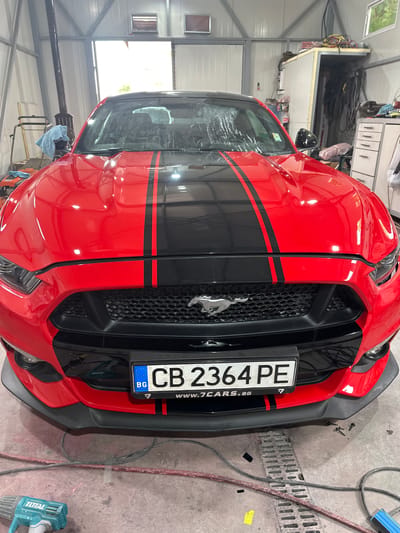 A vibrant red Ford Mustang sports car, adorned with bold black racing stripes down its hood and front bumper, is the central subject of the image. The car features a prominent black grille with a silver Mustang horse emblem. A Bulgarian license plate, displaying "CB 2364 PE," "BG," and the EU stars, is attached to the front bumper, along with "WWW.7CARS.BG" on its holder, indicating the car's location in Bulgaria.
The scene unfolds inside what appears to be a car workshop or garage. The setting is industrial, characterized by a concrete floor with a drainage grate, white corrugated metal walls, and exposed structural elements. Various tools and equipment are present; a blue "TOTAL" branded heat gun lies on the floor to the right of the car, and several screws are scattered near the car's front right side. Cables are sprawled across the floor and hung on the walls. In the background, storage shelves with items and what looks like a lifting hoist are visible.
Natural light filters in through windows, suggesting it is daytime. The car's red paint reflects the workshop environment, and its windshield exhibits streaks or water spots. No people are visible, and there are no discernible emotions. The car appears to be stationary, possibly undergoing or awaiting maintenance, detailing, or repairs within the workshop.