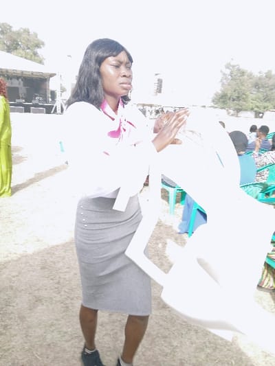 A woman stands outdoors in a white shirt, pink necktie, and grey skirt. She holds up a white plastic chair in front of her. Other people are seated in the background in blue and green chairs. The ground is a mix of green grass and dry dirt. There's a structure behind her that appears to be a building or a temporary stage.