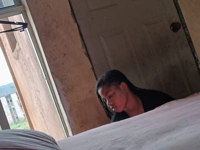 A young woman with braided hair and dark clothing is sitting on a bed with white sheets. She is looking down, seemingly lost in thought. Behind her is a weathered white door with a dark doorknob. To her left is a window with a black hanger visible. Through the window, a building with a light gray roof and visible windows is seen. Greenery is visible in the foreground outside the window. The room appears to be in a state of disrepair, with faded, stained walls. The image suggests an indoor setting in a room in Umueshi, Nigeria, with a woman sitting on a bed near a window and a door.