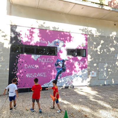 This image depicts a mural on the exterior wall of a building in Zaragoza, Spain. The mural is predominantly pink and features text in white and blue. Visible text includes "www.dazartses", "INGLÉS", "DANZA", and "MUSIDANZE", along with "TEATRO". A stylized depiction of the United Kingdom's flag is integrated into a brick pattern on the wall. A central element of the mural is a blue-clad figure in a dynamic dancing pose. Two young boys are in the foreground, facing away from the camera. The boy on the left wears a white t-shirt and blue shorts, while the boy on the right wears a red t-shirt with yellow trim and grey shorts. The ground is paved with small stones, and shadows from trees or overhead structures create a dappled effect on the scene. The building appears to be a modern structure, with visible windows and a light-colored facade. A small white electrical box with a red label is mounted on the adjacent wall.