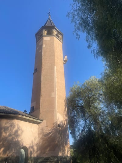 This image captures a close-up, low-angle view of a tall, tan-colored tower structure against a clear, bright blue sky. The tower is octagonal in shape and has a pointed roof, with decorative arches near the top. A thin, dark spire with some intricate metalwork extends from the very peak of the roof. The tower appears to be made of a stucco-like material, and there are some visible patches of lighter color that might indicate repairs or weathering. The surface of the tower is partially shaded by the leaves of large trees that arch over the top of the frame, suggesting it's a sunny day.
The surrounding environment shows glimpses of other architectural elements. A textured, dark-tiled roof or overhang is visible in the lower right corner, attached to a darker stone wall. To the left of the tower, a lighter, more modern-looking wall or building section is partially visible, with what looks like a satellite dish mounted on its upper edge. Wires run along the exterior of the tower, and a small, dark light fixture is attached to its side. The overall impression is of an older, possibly historical building complex set within a tree-lined area. The bright sunlight and shadows indicate it is daytime, likely midday or early afternoon. The image is taken from a ground-level perspective, looking upwards.
