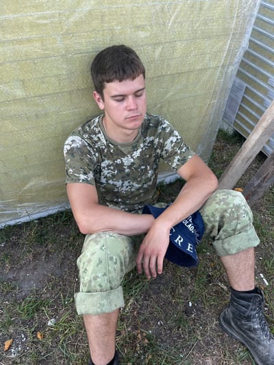 A young man in camouflage clothing sits with his eyes closed against a wall of yellow insulation panels. He is wearing a camouflage t-shirt and pants, with the pant legs rolled up above his knees. A blue bucket hat with white lettering is on his lap, obscuring part of his legs. His left arm is resting on his knee, and his right arm is crossed over his chest. The ground is covered in dirt and dry grass. To the right of the man, there are stacks of construction materials and wooden planks. The overall scene suggests a pause or break from outdoor work or activity. The image was taken in the Аснежыцкі сельскі Савет, Belarus.