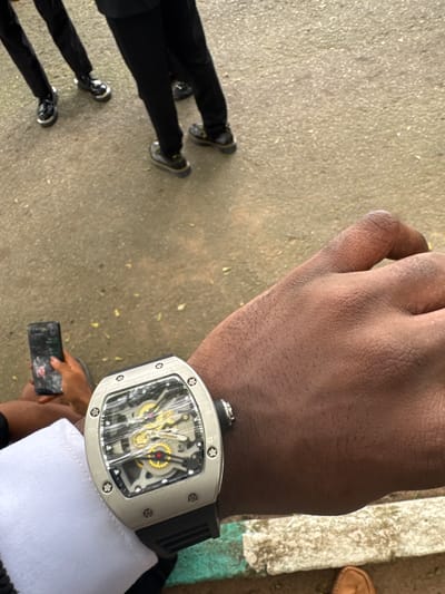 The image shows a man's wrist with a silver-cased watch. The watch has a skeletonized face with visible gears. He is wearing a white shirt with a black jacket. He is holding a smartphone. There are shoes and pants legs of multiple people visible on the left side of the image. The setting appears to be outdoors. The location is Eziobo, Nigeria.