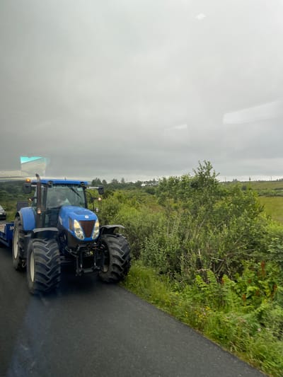 This image, taken in Cearnóg Sheain Bháin, Ireland, features a blue New Holland tractor parked on a wet asphalt road. The tractor is the primary subject, with its large, rugged tires and prominent headlights visible. The scene is set outdoors, with a blurred background of lush green fields and trees, suggesting a rural or agricultural environment. The sky is overcast and a bit hazy, indicating potentially damp or foggy weather.
The tractor appears to be stationary, and no specific activity is clearly depicted beyond its presence on the road. Notable details include the reflective surface of the road, which suggests recent rain or moisture. The overall impression is one of a practical, perhaps working, vehicle in a natural, somewhat muted setting.
The image has been rotated, making the ground appear on the right and the sky on the left. A slight reflection is visible on the left side of the frame, possibly from a window or screen through which the photo was taken. There is some faint text visible on the side of the tractor, but it is not legible. The time of day appears to be daytime, given the ambient light, but the overcast conditions obscure any direct sunlight. The mood of the image is neutral, focusing on the industrial subject within its natural surroundings.