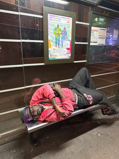A Black person lies on a bench at a bus stop in Garges-lès-Gonesse, France. The person, wearing a red jacket, jeans with floral embroidery, and glasses, is holding a phone. A poster is on the wall of the bus shelter, advertising "Les Rencontres de la Petite Enfance et de la Parentalité," an event from April 7 to 18, 2026. Another sign indicates bus line 9509 to Gare Dermont-Eaubonne. The ground is concrete.