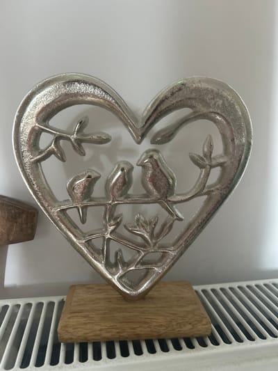 The image shows a silver heart-shaped decorative piece with three birds perched on a branch, all mounted on a wooden base. The piece is positioned in front of a white wall, with a white radiator visible in the foreground. To the left of the heart, a portion of another wooden object is visible. The lighting appears to be artificial, casting a slight shadow behind the decorative piece. The scene is an indoor setting, likely a home, and there is no information to suggest it is located in Limerick, Ireland. There are no people or actions depicted.