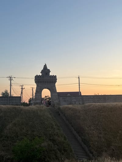 An outdoor, low-angle, full-frame shot depicts a concrete archway resembling a castle turret, set against a clear sky with a gradient of pale blue to warm orange hues, suggesting either sunrise or sunset. The archway stands on a grassy, elevated mound, accessible by a set of stone stairs. Several people, appearing to be a group of tourists, are visible near the top of the stairs and at the base of the archway, some standing and others seated. The background features a concrete wall, power lines, and utility poles, indicative of an urban or semi-urban setting. The overall scene conveys a sense of historical or memorial architecture integrated into a natural landscape.