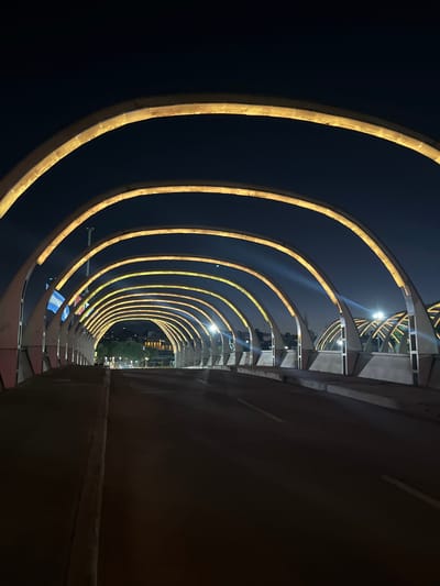 The image shows the interior of the Puente del Bicentenario, located in Cordoba, Argentina, at night. The perspective is from within the tunnel formed by the bridge's arches. Illuminated golden arches extend into the distance, diminishing in size. The road surface is visible to the right, with a center dividing line. The surrounding environment is dark, but distant city lights are visible at the tunnel's end. The bridge's structural elements are visible along the right side.