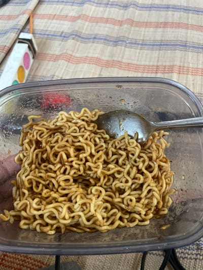 A close-up image depicts a clear plastic rectangular food container holding cooked instant noodles coated in a brown sauce, consistent with Indonesian instant fried noodles (Mie Goreng). A silver spoon rests within the noodles. Condensation is visible on the interior walls of the container, indicating the noodles are warm. The container is situated on a light-colored fabric surface featuring horizontal stripes in beige, blue, and reddish-orange. To the upper left, a portion of a white rectangular object with pink, yellow, and green circular markings is visible, with a thin yellow or orange cord extending upwards from it. A dark object is partially visible beneath the container. The scene is located in Kumbang, Indonesia. No individuals are present in the frame.