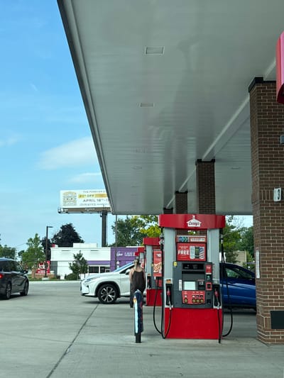 The image shows a gas station with red and white gas pumps under a white awning. A person is standing next to a pump, facing away from the camera. A white Lexus SUV is parked at a pump to the left of the Casey's branded pump. A black SUV is visible in the background to the left. A blue sedan is parked behind the Casey's pump. In the distance, there is a billboard with text and an image of jewelry. A sign that reads "Open LATE N" is visible on a building in the background. The gas station is located in Naperville, United States of America.