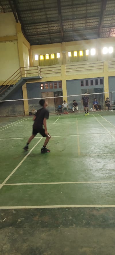 The image depicts an indoor badminton court with a green floor marked with white lines. In the foreground, a young male athlete in a black t-shirt and black shorts is in motion, playing badminton. His body is blurred due to movement, but his stance and the position of his racket suggest he is mid-swing or preparing to hit the shuttlecock. In the background, several other individuals are seated on chairs near the court, observing the game. One male athlete, wearing a dark t-shirt with a pattern and light-colored shorts, is also on the court, positioned further back and facing away from the camera. The court is enclosed by walls, with a net stretched across the middle. The ceiling appears to be made of corrugated metal, supported by visible beams. Natural light filters in through windows with yellow curtains along the upper walls. There is a set of stairs with a railing on the left side of the image, leading up to a higher level. The environment suggests a community sports facility, likely in Lhoksukon, Indonesia, given the context.