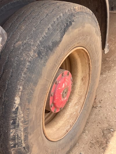 The image shows a close-up of a worn tire, likely from a vehicle such as a truck or tractor, resting on the ground. The tire is dirty and appears to have seen considerable use, with visible wear on the tread. Inside the rim of the tire, a red, circular hubcap with visible bolts is present. The background is a blurred expanse of dirt and small rocks, consistent with an outdoor, potentially rural or industrial setting. There are no people visible in the image. The lighting suggests it is daytime, but the weather is not discernible. Text is visible on the tire's sidewall, though it is partially obscured by dirt and the angle of the shot. The overall impression is one of a used, functional piece of equipment in a somewhat unkempt environment. The location context of Unguwar Saya, Nigeria, suggests this could be a scene related to transportation or agriculture in that region.