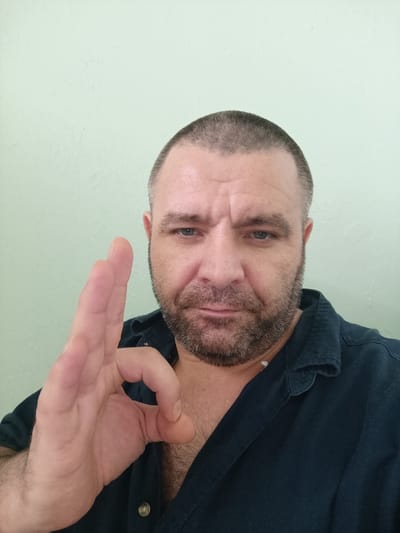 A medium shot shows a man with a shaved head and a beard, holding his right hand up in an "OK" gesture. He is looking directly at the camera with a serious expression. He is wearing a dark blue collared shirt. The background is a plain, light green wall. There is no visible indication of the location being Marshyntsi, Ukraine.