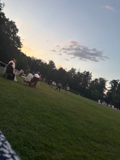 The image depicts a grassy field in Beltershain, Germany, during sunset. A group of people are scattered across the field, some sitting on chairs in the foreground and others engaged in activities further back. The sky is a blend of pale blue and orange hues, with clouds scattered across it. Trees line the background, forming a natural boundary to the field. The overall atmosphere suggests a casual outdoor gathering or event.