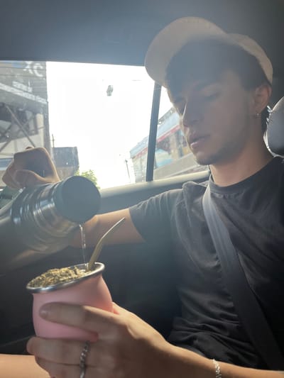 A young male, approximately 18-25 years old, is depicted seated in the rear passenger area of a vehicle in Hurlingham, Argentina, during daytime. He wears a light-colored baseball cap, a dark t-shirt, and a seatbelt across his torso. His facial features include light stubble and a labret piercing, with a silver earring visible in his left ear. A silver chain bracelet is present on his right wrist and a silver ring on his right hand. The individual is engaged in pouring water from a silver thermos, held in his left hand, into a pink mate gourd containing yerba mate and a bombilla, which he holds in his right hand. The vehicle's window displays an exterior street scene with bright sunlight, revealing distant buildings, power lines, and a partially visible red and white structure.