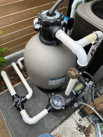 This image shows a pool pump and filter system set up outdoors. The main subjects are the pool equipment, including a large, beige sand filter tank, a black pump with a clear lid, and a complex network of white PVC pipes connecting them. The system is connected to a larger piece of equipment with a fan, possibly a pool heater or air conditioner unit, which is partially visible in the upper left. The equipment is situated on a paved surface, likely a patio or deck, with a wooden fence visible on the left and behind the equipment. Some fallen leaves and debris are scattered around the base of the pool filter.
There is no explicit activity or event taking place; the image appears to be a static shot of the pool equipment.
Notable details include the various gauges and labels on the equipment. The sand filter has a label that appears to say "HAYWARD". There is also a pressure gauge attached to the filter, which is difficult to read clearly. The pump has a label that says "HAYWARD Super II". The multi-port valve on the piping system has settings like "CLOSED" and "OPEN" visible. The time of day appears to be daytime, with natural light illuminating the scene. The weather is not definitively indicated but the presence of fallen leaves suggests it might be autumn. The location is identified as Mississauga, Canada. No people are clearly visible in the image, though a foot wearing a sandal is present in the bottom right corner. There are no discernible emotions conveyed by the image.
