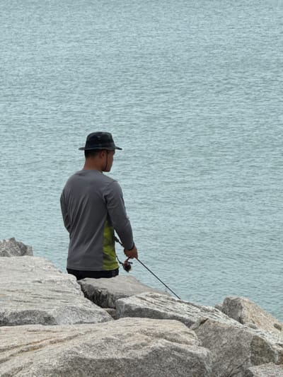 A person stands on rocky terrain, fishing with a rod and reel in hand. They wear a gray long-sleeved shirt, dark pants, and a hat. The body of water, with textured surface, extends to the left. The rocks are gray and craggy, taking up the right side of the image. The perspective is slightly tilted.