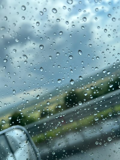 The image captures a close-up view of raindrops on a glass surface, likely a window. The raindrops are of various sizes and are scattered across the frame, creating a textured and reflective effect. Through the water droplets, a blurred background is visible, suggesting an outdoor scene. The background appears to be a mix of muted blues and grays, indicative of a cloudy sky, and streaks of green and gray, possibly representing foliage or a road with vehicles. The lighting suggests it is daytime, with soft, diffused light, consistent with overcast or rainy weather. The focus is entirely on the water droplets, which magnify and distort the view of the background, creating an abstract and atmospheric quality. There is no visible text in the image. The scene evokes a sense of a damp, possibly melancholic, day, common in a location like Vakarel, Bulgaria, during a period of rain.