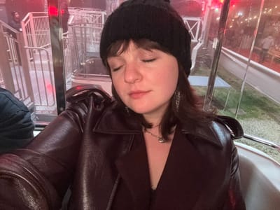 A person with closed eyes wears a black beanie and a dark brown leather jacket. They are positioned indoors, possibly within a glass or transparent structure, suggested by the visible metal railings and indistinct backgrounds. The scene outside shows blurry signs and faint lights, indicating an urban or entertainment district. The image appears to be taken at an angle, possibly from a slightly elevated position.