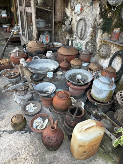 The image depicts an outdoor display of various traditional kitchenware and decorative items, likely for sale or as part of a cultural exhibit. The scene is set against a rustic, textured wall with patches of moss and grime, suggesting an aged outdoor location. In the foreground, a collection of clay pots of different sizes and shapes are arranged on a textured concrete surface, some with lids, others open. Several metal cooking vessels, including large woks and smaller pots, are also present. A rolling pin rests on a rustic wooden stand, alongside a small wire basket. Other objects include a tin kettle, a beige plastic jerrycan, and smaller ceramic items like bowls and cups. A wooden cabinet with wire mesh doors stands in the background, holding more ceramic plates and pots. The overall impression is one of a storage or display area filled with handcrafted, possibly antique, items. The location is identified as Banguntapan, Indonesia, known for its crafts and cultural heritage.