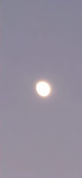 This vertically oriented photograph, captured in the city of Vendin-le-Vieil, France, depicts a singular, prominent celestial body or light source against a uniformly featureless sky. The central object is a bright, circular emission of white-yellow light, surrounded by a soft, luminous halo. The sky maintains a consistent muted grey-purple hue, indicating either heavy overcast conditions, twilight, or significant atmospheric haze. There are no clouds, other celestial phenomena, ground features, artificial structures, or human presence discernible within the frame. The scene is static, devoid of any discernible actions or interactions, focusing solely on the atmospheric light phenomenon.