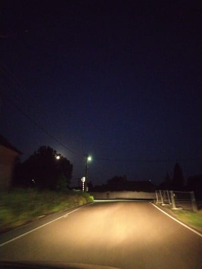 This is a nighttime, low-light, interior shot from a vehicle moving along a road in Veselá, Czechia. The road ahead is illuminated by the vehicle's headlights, casting a warm glow on the asphalt. A white line delineates the right edge of the road. To the right of the road, a grassy embankment rises, topped with some vegetation and a modern fence structure. Beyond the fence, the silhouette of trees and buildings is visible. To the left of the road, there is more vegetation and the dark shape of trees. A streetlight stands on the right side of the road in the middle ground, and a power line stretches across the dark sky. The sky is a deep blue-black, with a faint appearance of a crescent moon or a distant light source. The overall impression is one of solitary travel on a rural road after dark.