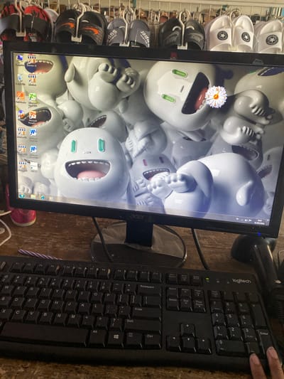 The image shows a desktop computer monitor displaying a repeating pattern of cartoon-like, white, open-mouthed characters. A single white flower is digitally superimposed on one of the characters. The monitor is sitting on a brown wooden surface alongside a black Logitech keyboard, with a person's finger visible near the numpad enter key. The computer's taskbar displays several application icons, including Recycle Bin, Foxit Reader, Excel 2010, PowerPoint, and Microsoft Word. To the left of the monitor, a shoe rack is visible, filled with various sandals and slides, including brands like Converse and Nike. A purple and white pen lies next to the keyboard. The monitor displays a date of 31/05/2026. The image was taken indoors in Mojogedang, Indonesia.