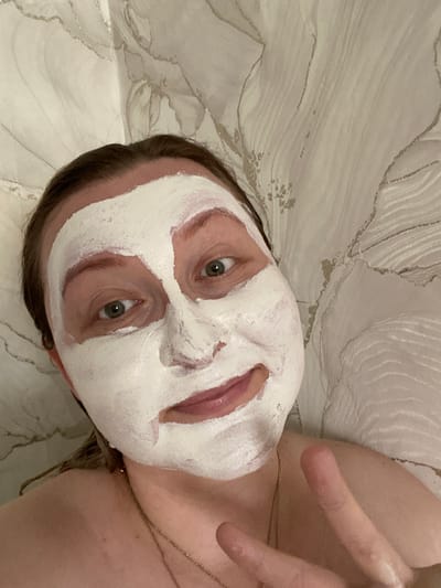 A woman with a white face mask applied to her face is taking a selfie. She has medium-length brown hair and is wearing a necklace. She is making a peace sign with her right hand. The background appears to be a wall with a textured pattern. The image was taken in Oktyabrsky, Russia.