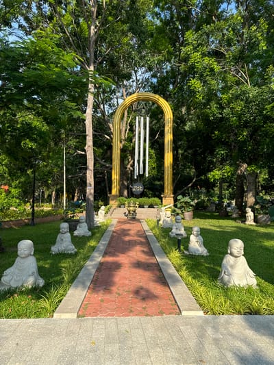 The image depicts a tranquil garden setting with a pathway leading towards a golden archway. The pathway is paved with red bricks, bordered by lush green grass and ornamental plants. Along the pathway, several stone statues of young Buddhist monks are strategically placed, each in a seated meditative pose. These statues add to the serene and spiritual ambiance of the garden.

In the background, a large golden arch stands prominently, adorned with wind chimes. The arch is supported by two fluted columns and is framed by mature trees with dense green foliage. The sunlight casts shadows on the pathway, indicating it is likely daytime, possibly late afternoon given the warm tones. The overall scene suggests a well-maintained garden, possibly part of a temple or a place of spiritual retreat. The presence of the statues and the archway implies a religious or cultural significance to the location. The environment is natural and peaceful, with the vibrant green of the trees and grass contrasting with the golden arch and red pathway.