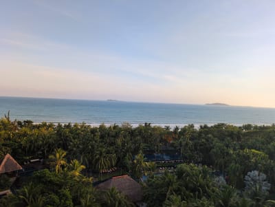 The image captures an aerial view of a tropical resort area in Tianya District, China, at sunset. The scene is dominated by lush green palm trees and dense foliage, interspersed with swimming pools and thatched-roof structures. In the distance, the tranquil sea meets a clear, pale blue sky, with two small islands visible on the horizon. The overall ambiance is one of serenity and natural beauty.