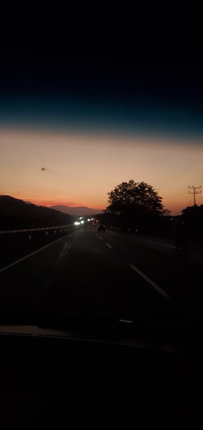The image depicts a highway at dusk, likely near San Joaquín, Venezuela, based on the tags. The sky transitions from dark blue to orange and pink near the horizon, suggesting a sunset or sunrise. Silhouette of mountains and power lines are visible in the background. A tree stands beside the road. Cars with headlights on are seen on the highway. The highway itself is dark with visible lane markings.