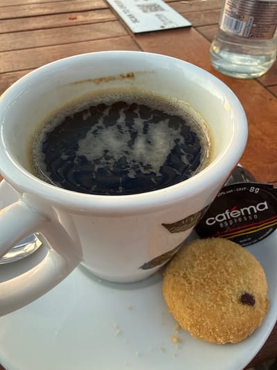 A close-up, eye-level shot shows a white coffee cup filled with dark coffee, resting on a white saucer. A small, round, golden-brown cookie with a hint of chocolate is placed on the saucer to the right of the cup. A dark, rectangular packet with the word "cafema espresso" printed on it is positioned behind the cookie and to the right of the cup. The scene is set on a slatted wooden table. In the background, out of focus, is a clear glass bottle of water and a white card with some text and a QR code on it. The lighting suggests it is daytime, possibly late afternoon or early evening, with warm tones illuminating the scene. This image was taken at the beach of Hook of Holland, Netherlands.