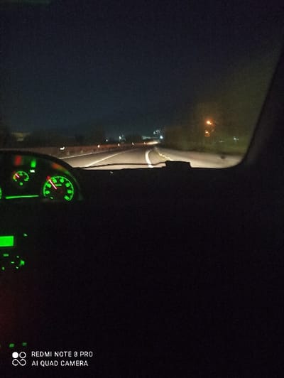 The image depicts a nighttime view from inside a vehicle, reportedly located in San Valentino Torio, Italy. The perspective is forward through the windshield, capturing a section of road illuminated by the vehicle's headlights.
The lower portion of the image shows the vehicle's dashboard, specifically the left side where the instrument cluster is visible, glowing with green illumination. Two primary circular gauges and other smaller indicators or displays are discernible within the cluster.
The road ahead features white solid and dashed lane markings. To the immediate left of the road, a roadside barrier, possibly a guardrail, is visible with reflective elements. Further along the left side, distant artificial lights suggest the presence of a town or populated area. To the right of the road, dark vegetation and a single streetlamp emitting an orange glow are present. No individuals are depicted within the scene. A "REDMI NOTE 8 PRO AI QUAD CAMERA" watermark is present in the lower-left corner of the image. The exact location within San Valentino Torio is not visually identifiable from the image content.