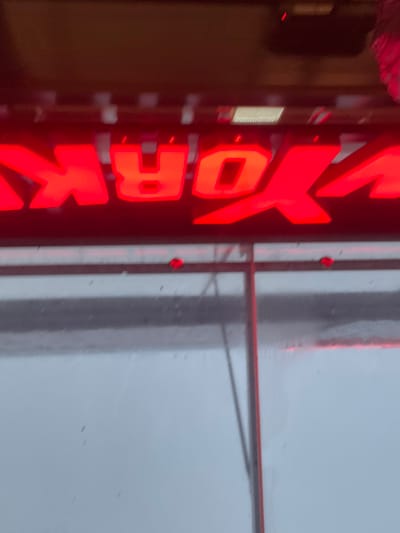 The image displays a red neon sign featuring the letters "YORK" prominently in the upper frame. A dark, horizontal structural beam is visible directly above the illuminated letters. Below the neon sign, a transparent panel, consistent with a window or glass barrier, occupies the central and lower portions of the image. This panel shows evidence of surface moisture, including visible droplets and smudges. Dark structural elements, specifically a vertical support and two diagonal braces, are discernible behind or within the transparent panel, indicating an internal framing system. The panel's surface reflects a blurry, diffuse light gray field, suggesting an overcast sky or an interior light source. The scene is located in the city of Reichstett, France, under conditions of low ambient light, with the red neon sign serving as the primary light source for the depicted objects.