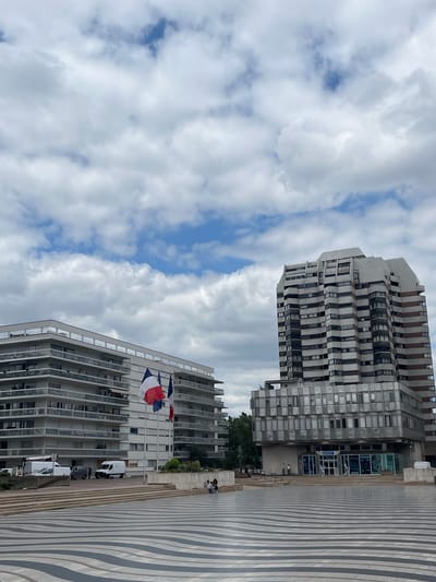 In the city of Créteil, France, a wide, black and white striped plaza occupies the foreground. Beyond the plaza, a modern, multi-story residential building with numerous balconies stands on the left. To the right, a taller, more angular building features a facade of windows and darker horizontal bands. Two French flags are prominently displayed on flagpoles in front of the residential building. Several white vans and one dark colored vehicle are parked near the entrance of the residential building. Further to the right, near the base of the taller building, several people are seated on a raised concrete area. A few individuals are also walking near the entrance of the taller building. The sky above is filled with a mix of white and gray clouds, with patches of blue sky visible.