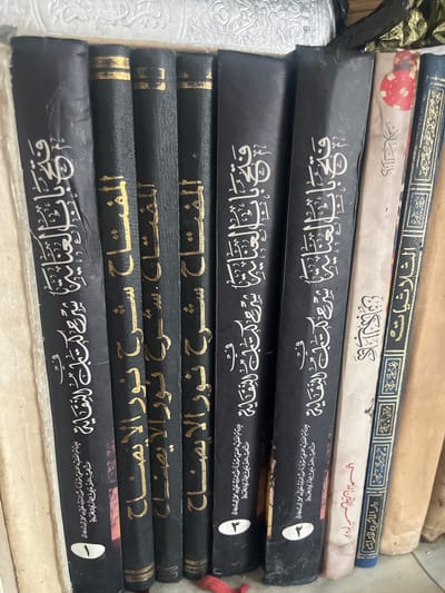 The image shows a close-up view of a bookshelf filled with books. The books are bound in dark covers, some with gold lettering, and others with white text. The spines of the books are visible, displaying Arabic script. The books are arranged vertically, standing upright on a shelf. The background is blurred, but it appears to be a domestic setting, possibly a home or a library. The lighting is natural, casting soft shadows on the books. The overall impression is one of a well-stocked collection of religious or scholarly texts. There is no information or context to indicate the location in the city of Ташлак, Kyrgyzstan.