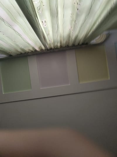 The image shows a close-up of a light green curtain with a subtle pattern of white figures and thin black wavy lines. The curtain is pleated and hangs to the left of the frame, obscuring a portion of the background. To the right of the curtain, there are three square panels on a light grey wall, arranged vertically. The top panel is a light yellow, the middle panel is a soft lavender, and the bottom panel is a pale green. The lighting suggests it is daytime, possibly indoors, with a soft, diffused light source. The overall impression is one of a calm and orderly domestic setting. There is no visible text in the image. The provided location context of Melitopol, Ukraine, does not offer further visual cues within the image itself.