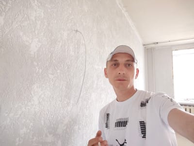 The image shows a man wearing a white Puma t-shirt and a white baseball cap, taking a selfie in what appears to be a room undergoing renovation or construction. The walls have a textured, off-white finish, and there is a faint, curved line drawn on the wall to the left of the man, possibly a marking for work. The ceiling is also visible, with some electrical conduit. To the right, a window with bright light coming through is partially visible, along with a radiator. The man is holding a cigarette in his right hand. The scene is indoors, likely residential, and the presence of construction materials or unfinished work suggests it could be in the city of Bor, Russia, as indicated by the context.